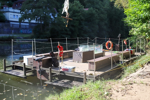 Das Floß liegt am Steg. Bänke, Aquarien, Kescher und weitere Utensilien sind zu sehen.