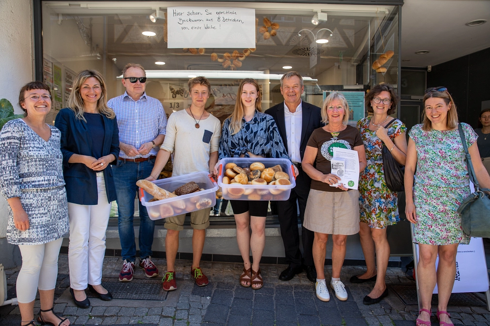 Vor der Schaufenster-Scheibe eines Ladens in der Marburger Oberstadt stehen mehrere Menschen, die in die Kamera lächeln. Zwei von ihnen halten durchsichtige Körbe mit Backwaren in den Händen. Über ihnen hängt ein Schild mit der Aufschrift "Hier sehen Sie gerettete Backwaren aus 8 Betrieben von einer Woche!"