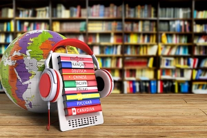 Fremdsprachen in der Stadtbücherei Vor mehreren Bücherregalen steht im Vordergrund auf einer Holzplatte eine Halterung mit mehreren Fremdsprachen-Büchern. Auf deren Buchrücken stehen die Worte "English", "Français", "Deutsch", "Chinese", "Italiano", "Polski", "Svenska", "русский" und "Canadian". Auf dem Gestell hängt ein rot-grauer Kopfhörer.