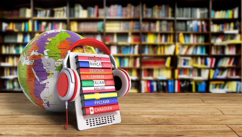 Fremdsprachen in der Stadtbücherei Vor mehreren Bücherregalen steht im Vordergrund auf einer Holzplatte eine Halterung mit mehreren Fremdsprachen-Büchern. Auf deren Buchrücken stehen die Worte "English", "Français", "Deutsch", "Chinese", "Italiano", "Polski", "Svenska", "русский" und "Canadian". Auf dem Gestell hängt ein rot-grauer Kopfhörer. © iStock/NosUA