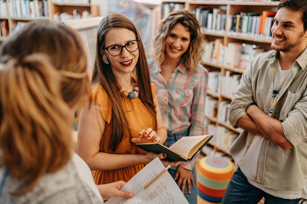 Führungen für Erwachsene in der Stadtbücherei Vor mehreren Bücherregalen steht eine vierköpfige Gruppe erwachsener Menschen. Der Fokus liegt auf der Frau in der Mitte, die ein aufgeschlagenes Buch in den Händen hält und etwas erzählt. Die Zuhörenden lächeln und schauen sie aufmerksam an. © iStock/StefaNikolic