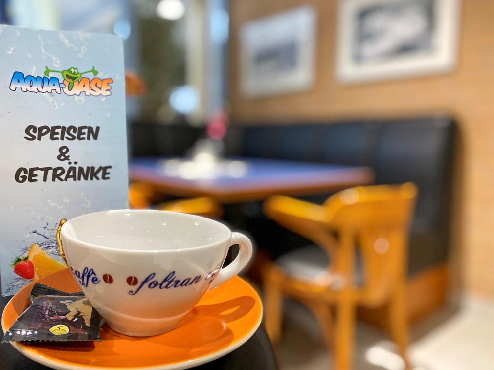 Gastronomie im AquaMar Eine Kaffeetasse auf einem Unterteller mit einem Keks, dahinter steht die Speisekarte mit der Aufschrift "AquaOase Speisen & Getränke", im Hintergrund ist eine Sitzeecke. © Simone Batz, Stadt Marburg