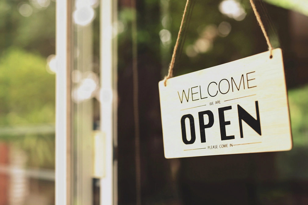 An einer Glasscheibe einer Eingangstür hängt ein Schild mit der Aufschrift "Welcome, we're open" ("Willkommen, wir haben geöffnet).