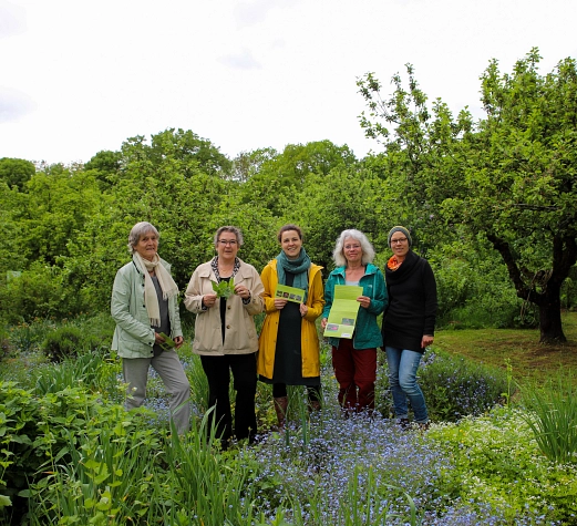 Gesundheitsgarten 2026 Fünf Frauen stehen in einem Garten, umringt von Büschen und Bäumen. In der Hand halten sie Flyer.