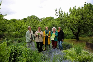 Gesundheitsgarten 2026 Fünf Frauen stehen in einem Garten, umringt von Büschen und Bäumen. In der Hand halten sie Flyer.