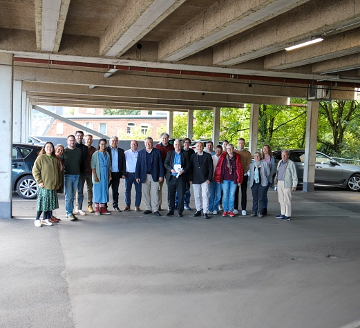 Eine Personengruppe steht auf einem Parkdeck. Im Hintergrund sind Autos zu sehen.