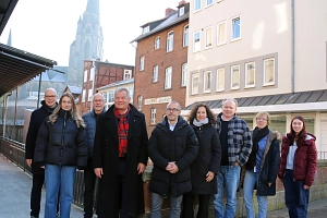 Eine Gruppe von Menschen steht in einer Straße. Im Hintergrund ist ein Kirchturm zu sehen.