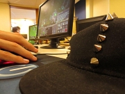 Eine Nahaufnahme zeigt eine Hand an einer Computermaus, im Hintergrund ein Monitor und vorne rechts eine Bascap.