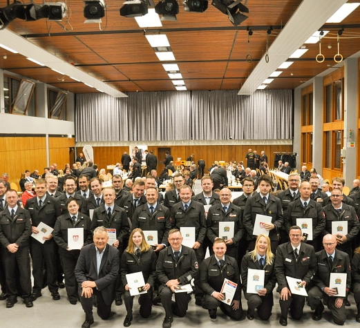 JHV 2026 FFW MR Eine große Gruppe von Feuerwehrfrauen und -männern stehen gemeinsam in einem Saal. Viele halten eine Urkunde in den Händen. Alle lächeln in die Kamera. Im Hintergrund sind weitere Personen zu sehen.