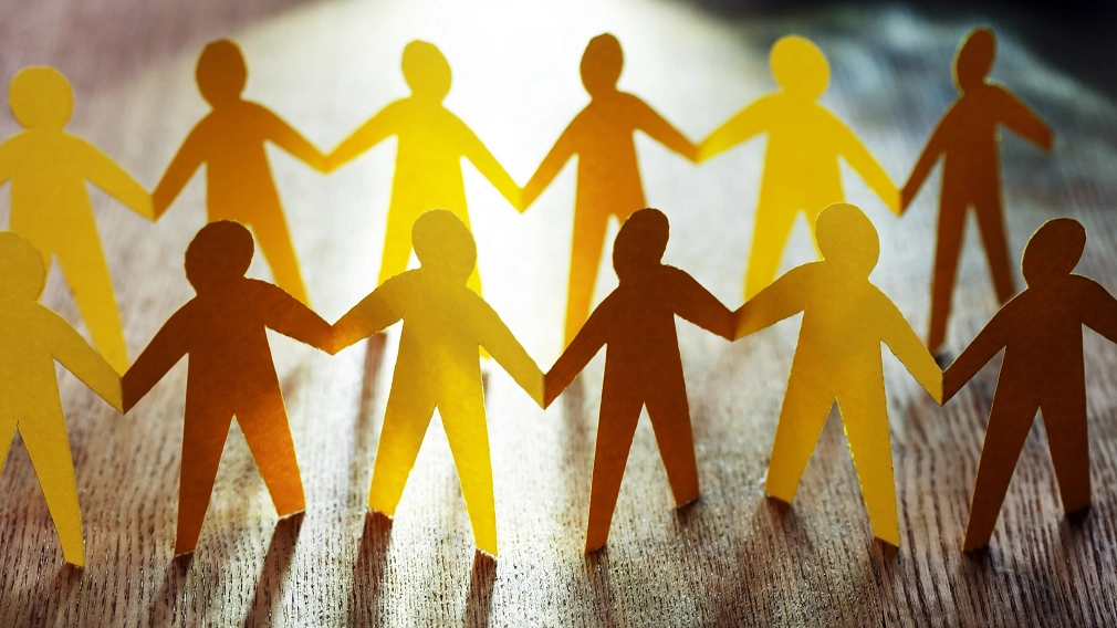 Kampagne gegen Einsamkeit: Gemeinsam statt einsam_iStock-1466755501_Ausschnitt.jpg Scherenschnitt aus gelbem Papier auf Holzuntergrund: Gruppe von Menschen, die sich an den Händen hält. © iStock/kvkirillov