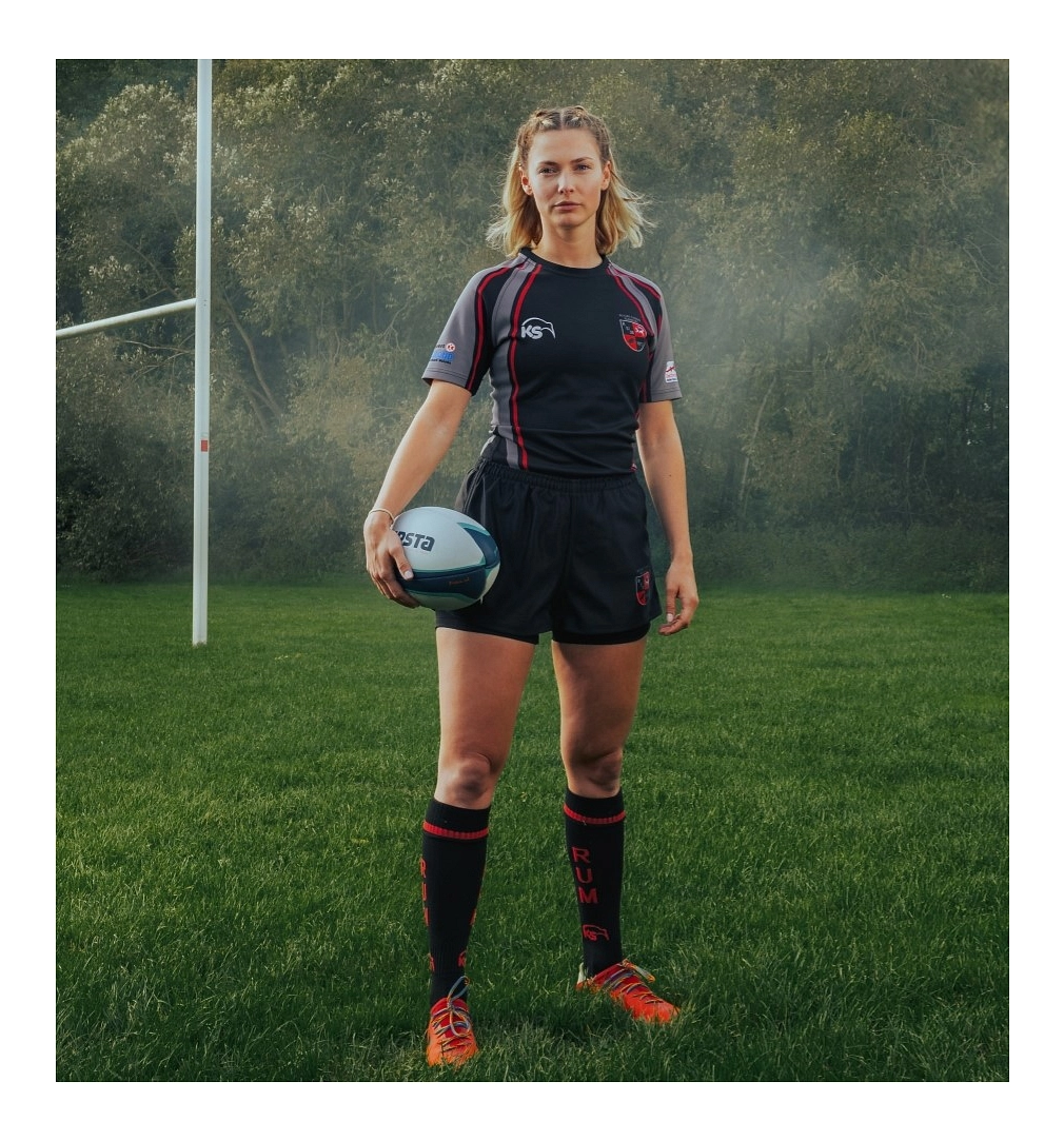 Eine Rugby-Spielerin steht auf dem Spielfeld. Blonde, schulterlange wellige Haare, Blick in die Kamera. Leichter Nebel.
