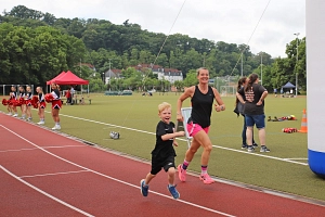 Kinder und Familientriathlon 2025 Eine Frau und ein kleines Kind laufen gemeinsam auf einer Laufbahn in einem Stadion. Im Hintergrund sind eine grüne Rasenfläche, einige Personen, ein Zelt und Bäume zu sehen.