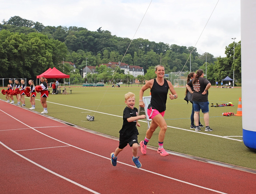 Eine Frau und ein kleines Kind laufen gemeinsam auf einer Laufbahn in einem Stadion. Im Hintergrund sind eine grüne Rasenfläche, einige Personen, ein Zelt und Bäume zu sehen.