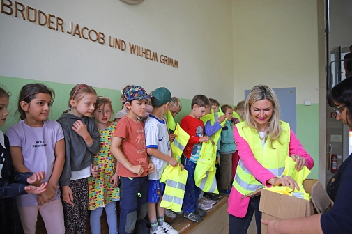 Das Bild zeigt eine Frau, die einer Gruppe von Kindern Warnwesten austeilt.