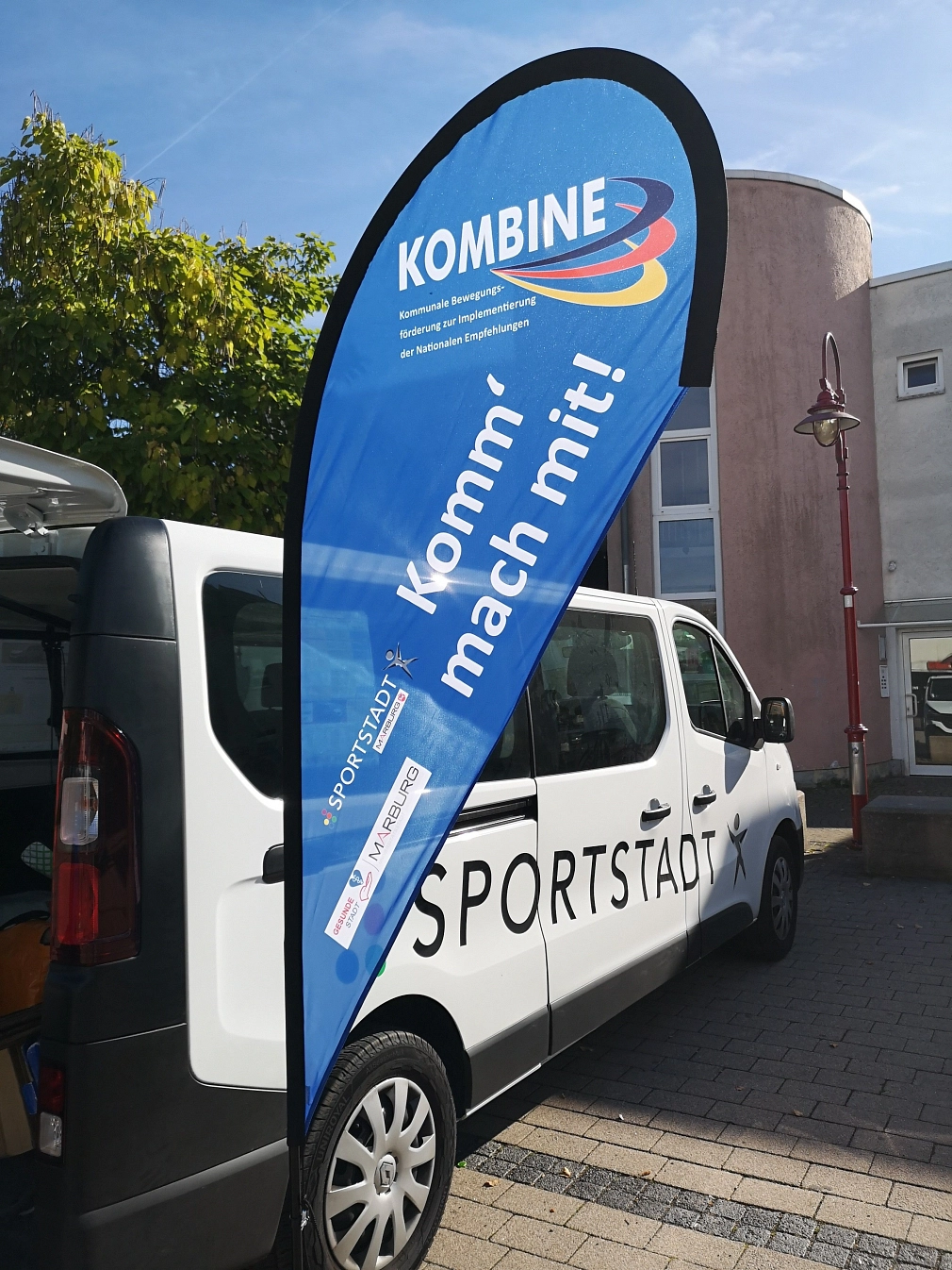 KOMBINE-Bus und Beach-Flag Ein weißer Kleinbus mit der Aufschrift "Sportstadt" sowie eine blaue Beach-Flag mit der Aufschrift "KOMBINE" und "Komm' mach mit!" stehen vor einem Gebäude. © Universitätsstadt Marburg