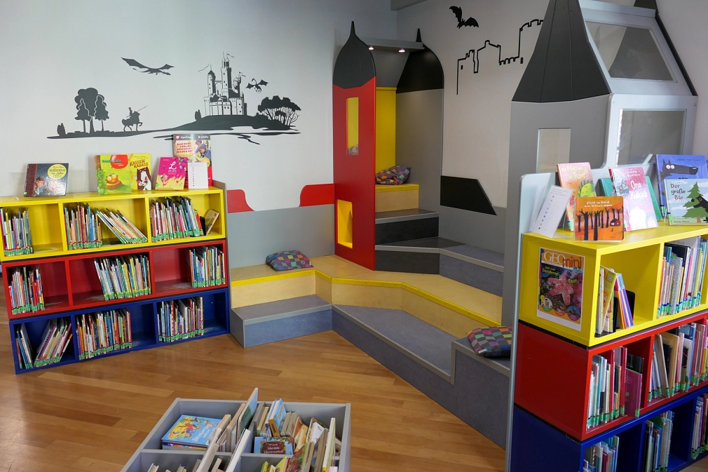 Leseburg der Kinderbücherei in der Stadtbücherei Ein Ausschnitt aus der Kinderbücherei in der Stadtbücherei: Die sogenannte Leseburg mit nachgebildeten Burgzinnen und Sitzmöglichkeiten, daneben gelbe, rote und blaue Bücherregale, an den Wänden Wandtattoos mit Drachen, Ritter und Burg © Universitätsstadt Marburg