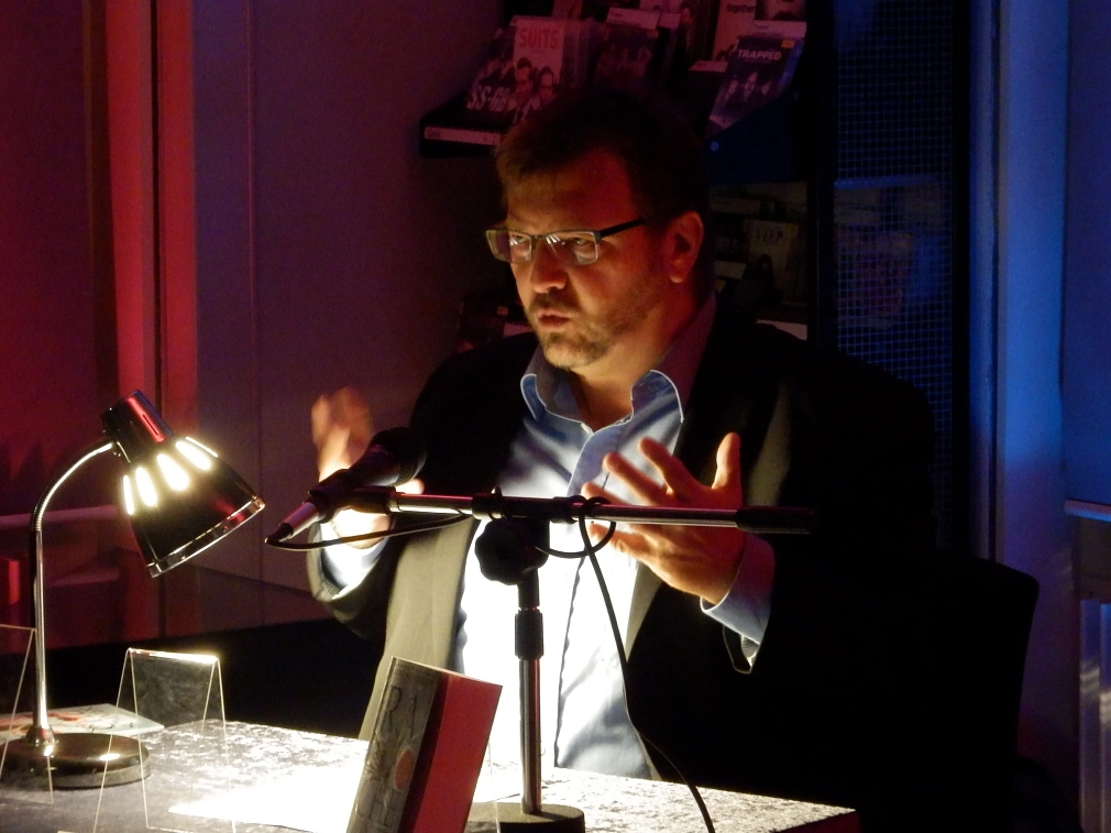 Ein Mann sitzt an einem Tisch, auf dem eine Lampe und ein Mikrophon stehen. Er gestikuliert und spricht.