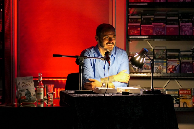 Der Schriftsteller Max Goldammer spricht in der Stadtbücherei in ein Mikrofon. Der Tisch, an dem er sitzt, ist spärlich beleuchtet. Im Hintergrund ist ein rotes Rollo heruntergelassen, DVDs stehen in den Regalen.&nbsp;&copy;&nbsp;Universitätsstadt Marburg