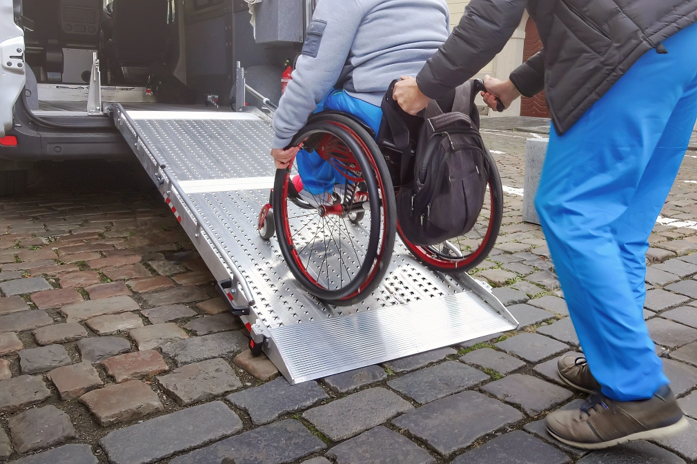 Marburg-Behindertenfahrdienst-iStock-1404919512 Eine Person schiebt eine andere Person im Rollstuhl auf einer barrierefreien Autorampe in ein Fahrzeug. © iStock / 24K-Production