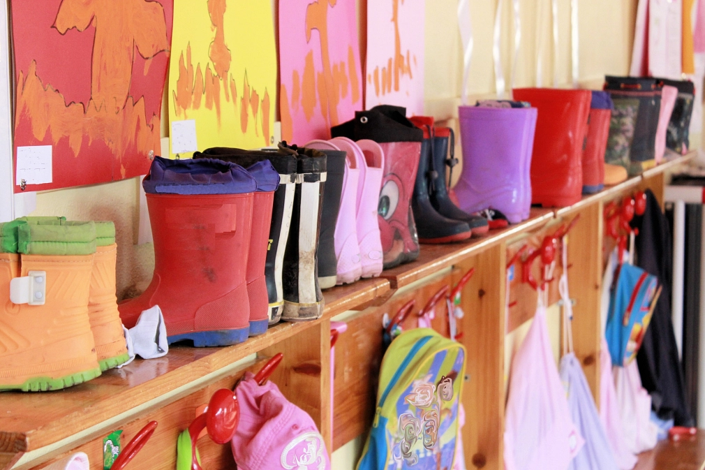 Garderobe eines Kindergartens mit bunten Gummistiefel und Haken mit Rucksäcken. Darüber an der Wand selbstgemalte Bilder.