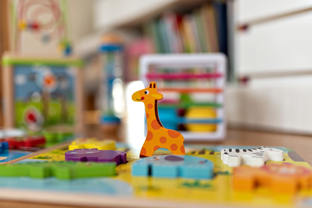 Tisch mit buntem Spielzeug, im Mittelpunkt steht eine kleine orangefarbene Spielzeuggiraffe aus Holz. Im Hintergrund sind unscharf weitere Spielzeugteile und Bücher zu erkennen.