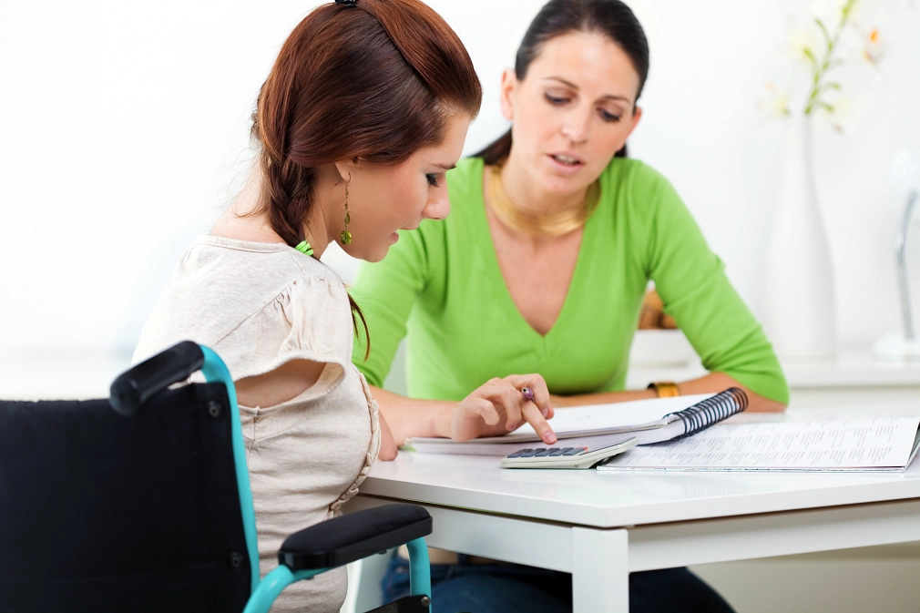 Eine junge Frau im Rollstuhl sitzt mit einer Beraterin am Tisch. Dort liegen ein Notizblock und ein Taschenrechner.&nbsp;&copy;&nbsp;iStock / izusek