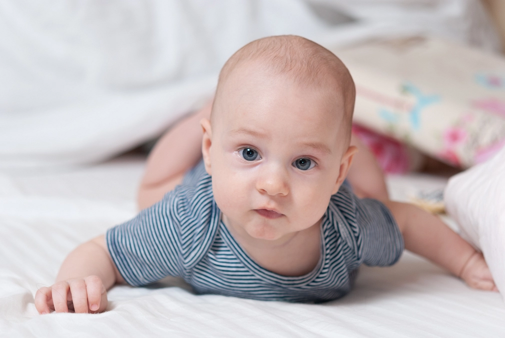 Ein vier Monate altes Baby liegt auf dem Bauch auf einem Bett. Es guckt in die Kamera und lächelt.&nbsp;&copy;&nbsp;iStock / Andrii Tolstopiatykh