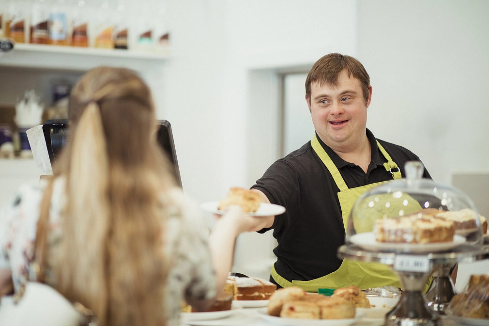Marburg-Menschen-Arbeit-Behinderung-iStock-1162977284 Ein Mann mit Down-Syndrom serviert einer Frau in einem Café, in dem er arbeitet, ein Stück Kuchen. © iStock / SolStock