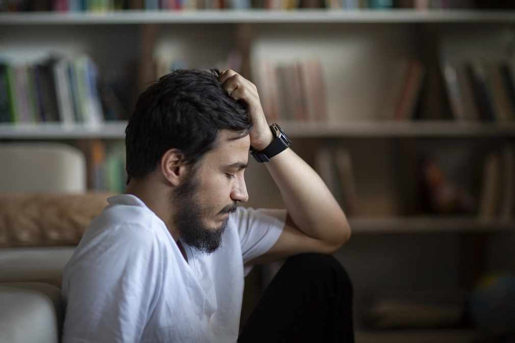 Ein junger Mann sitzt auf dem Sofa, die Augen geschlossen, der Kopf in die Hand gestützt. Erwirkt deprimiert. Im Hintergrund ein Bücherregal.&nbsp;&copy;&nbsp;iStock / ridvan_celik