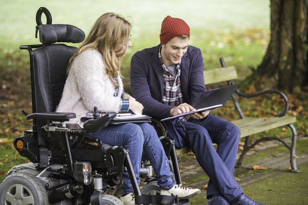 Marburg-Studierende-Behinderung-iStock-680248362 Eine junge Frau im Rollstuhl sitzt neben einem jungen Mann, der auf einer Parkbank sitzt, im Park. Er hält ein Tablet, beide unterhalten sich. © iStock / vm