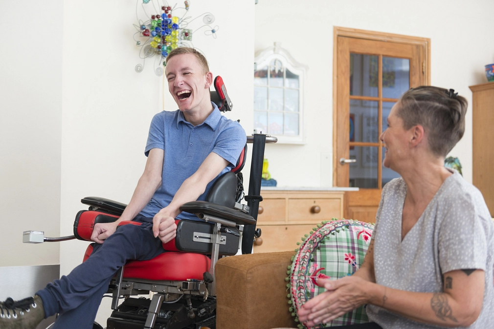 Marburg-Wohnen-Behinderung-iStock-603871162 Ein junger Mann im Rollstuhl und eine Frau, die auf einem Sofa sitzt, unterhalten sich. Beide lachen. © iStock / funky-data