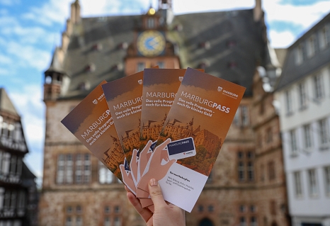 Flyer des MarburgPasses werden hochgehalten. Dahinter ist das Marburger Rathaus zu sehen. © Stefanie Ingwersen, Stadt Marburg