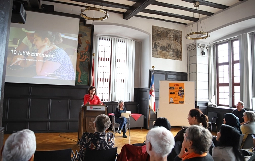 Eine Frau steht an einem Rednerpult. Im Hintergrund ist auf einer Leinwand eine Präsentation zu sehen.