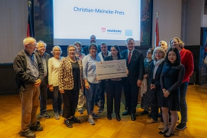 Meineke-Preis Das Bild zeigt viele Menschen nebeneinander, eine Frau hält einen großen Check der Sparkasse in der Hand. Auf einer großen Leinwand im Hintergrund steht "Christian-Meineke-Preis".