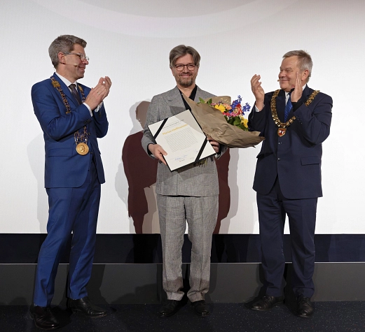 Drei Männer stehen auf einer Bühne. Der Mann in der Mitte hält einen Blumenstrauß und eine Urkunde in der Hand. Die beiden Männer links und rechts von ihm klatschen in die Hände.