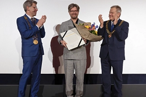 MR Kamerapreis 2026 Drei Männer stehen auf einer Bühne. Der Mann in der Mitte hält einen Blumenstrauß und eine Urkunde in der Hand. Die beiden Männer links und rechts von ihm klatschen in die Hände.