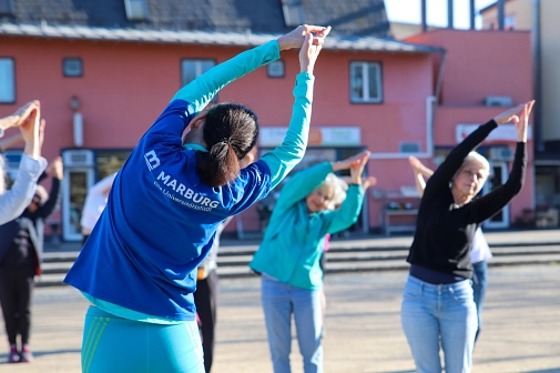 Mehrere Personen halten ihre Arme gestreckt über den Kopf, die Handflächen aneinander gelegt. Die Oberkörper sind zu einer Seite gebeugt.