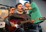 Ein Mann sitzt auf einem Frisörstuhl, singt und spielt Gitarre, während ihm die Haare geschnitten werden.
