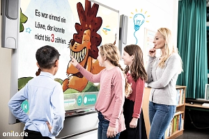 Drei Kinder und ihre Lehrerin stehen vor einem Whiteboard mit dem Bild eines lächelnden Löwen.