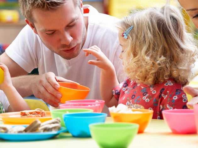 Essenszeit im Kindergarten: Junger Mann mit kleinen Kinder, vor ihnen auf dem Tisch stehen bunte Teller und Schälchen.