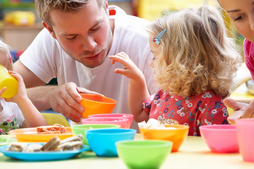 Essenszeit im Kindergarten: Junger Mann mit kleinen Kinder, vor ihnen auf dem Tisch stehen bunte Teller und Schälchen.