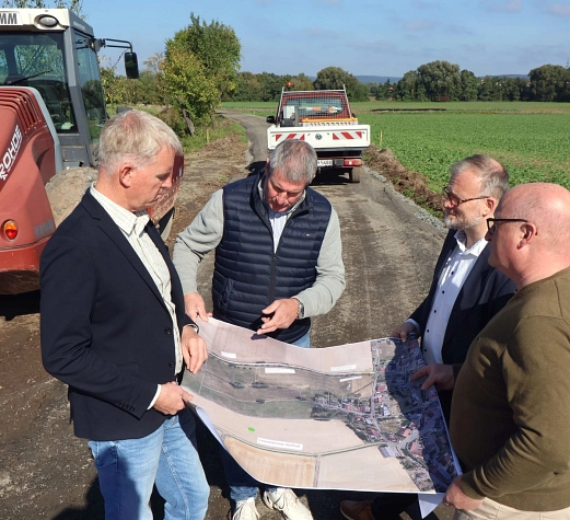 Vier Männer stehen auf einem Feld. Sie halten Baupläne in der Hand. im Hintergrund sind Baumaschinen zu sehen.