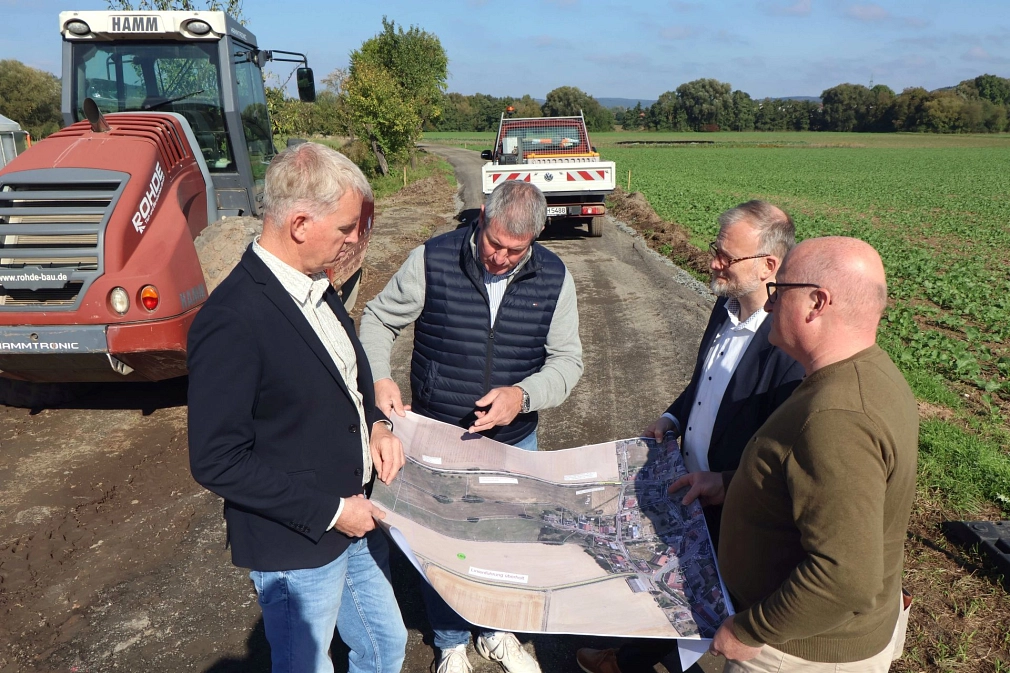 Vier Männer stehen auf einem Feld. Sie halten Baupläne in der Hand. im Hintergrund sind Baumaschinen zu sehen.
