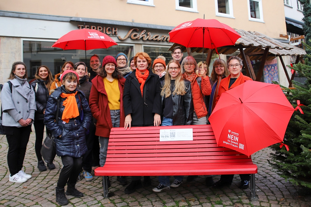 Das Bild zeigt eine Gruppe von Personen hinter der Roten Bank stehend, die in die Kamera lächeln. Drei Personen halten einen passenden roten Regenschirm.