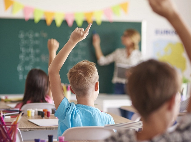 Kinder, die sich melden, sitzen in einem Klassenraum auf ihren Stühlen. Der Blick geht über ihre Köpfe hinweg nach vorne zur Tafel, wo die Lehrerin steht.
