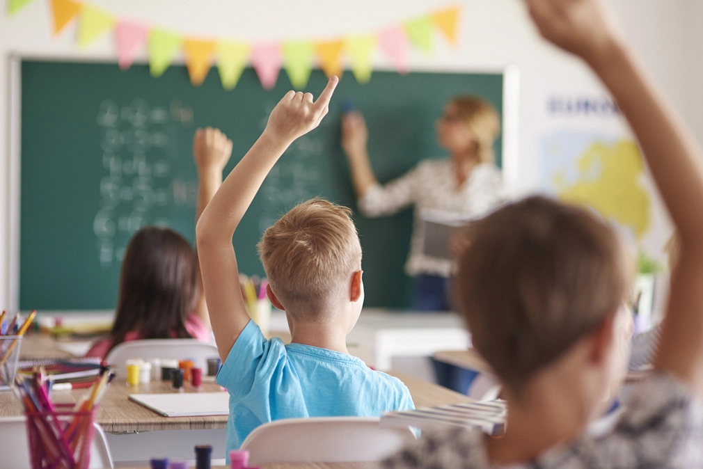 Rund um Schule Kinder, die sich melden, sitzen in einem Klassenraum auf ihren Stühlen. Der Blick geht über ihre Köpfe hinweg nach vorne zur Tafel, wo die Lehrerin steht. © Istock/gpointstudio