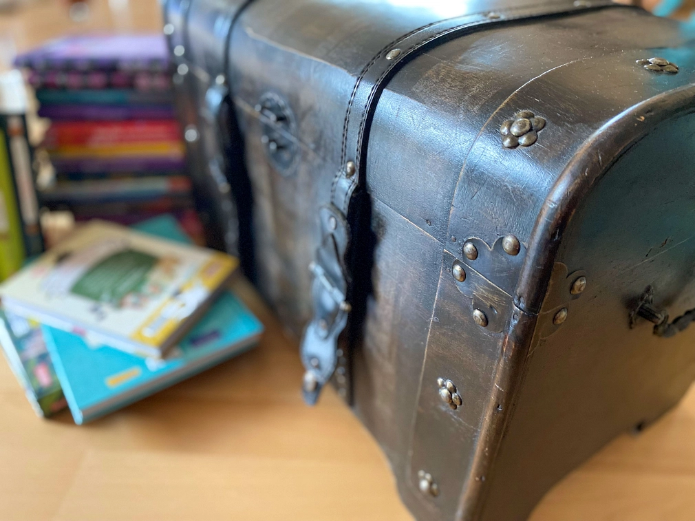 Schatzsuche mit Schatztruhe für Kinder in der Stadtbücherei Eine braune Truhe, die wie eine Schatzkiste aussieht, steht auf einem Holzbuden. Im Hintergrund und davor liegen gestapelt mehrere Kinderbücher. © Simone Batz, Stadt Marburg