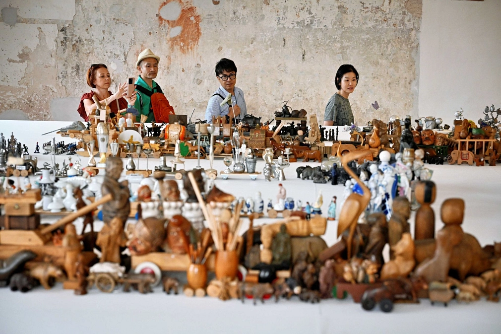 Kleine Figuren aus Holz und anderen Materialien stehen auf Tischen. Vier Besucher schauen sich diese Figuren an.