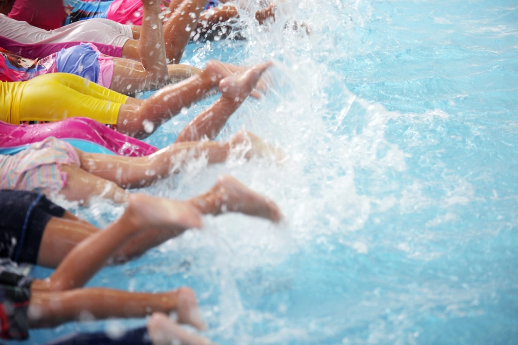 Schwimmkurse für Kinder im AquaMar und im Hallenbad Wehrda Eine Gruppe von Kindern liegt auf dem Bauch am Beckenrand und lässt die Beine auf- und abwärts ins Wasser schnellen, so dass das Wasser hochspritzt. Die Köpfe sind nicht zu sehen. © iStock/sakhorn38