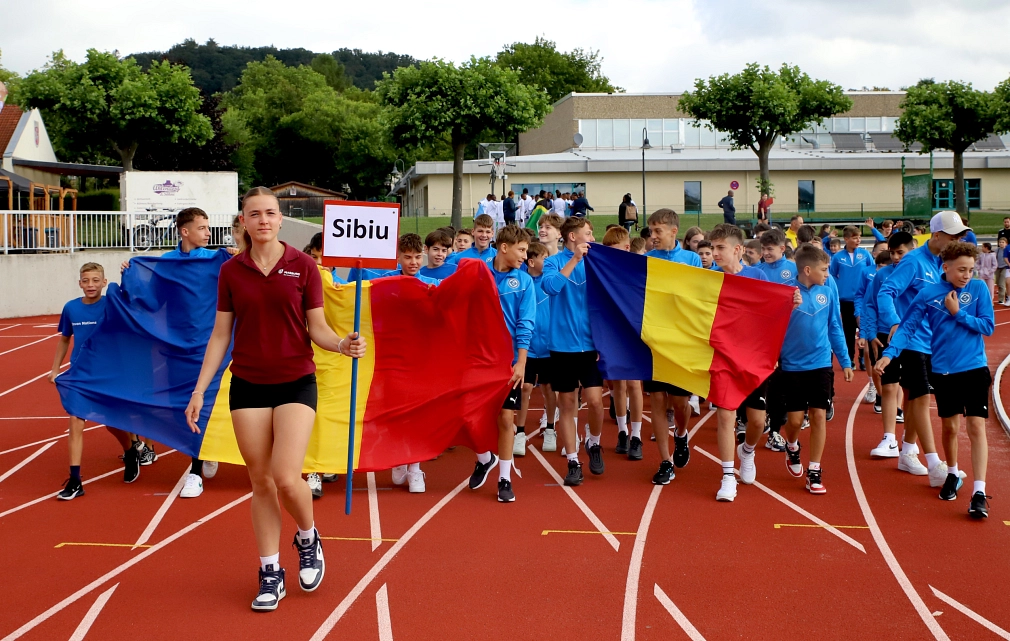 Ein Mädchen aus dem Seven Nations Camp 2024-Team trägt ein Schild mit der Aufschrift "Sibiu", hinter ihr laufen Jungen mit blauen Oberteilen, die  rumänische Flaggen tragen. Sie laufen gemeinsam ins Georg-Gassmann-Stadion ein.